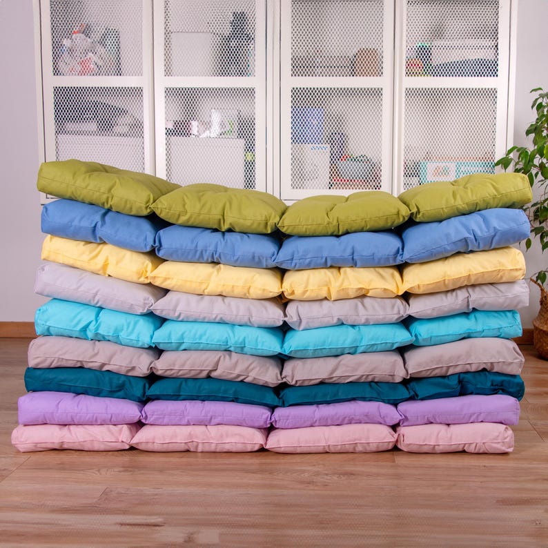 May include: A stack of 15 colorful cushions in various shades of green, blue, yellow, turquoise, gray, teal, purple, and pink. The cushions are arranged in a neat pile, with the top cushion being green.