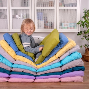 May include: A child sits on a stack of colorful cushions, arranged in a zig-zag pattern. The cushions are made of soft fabric and are perfect for creating a cozy and comfortable seating area.