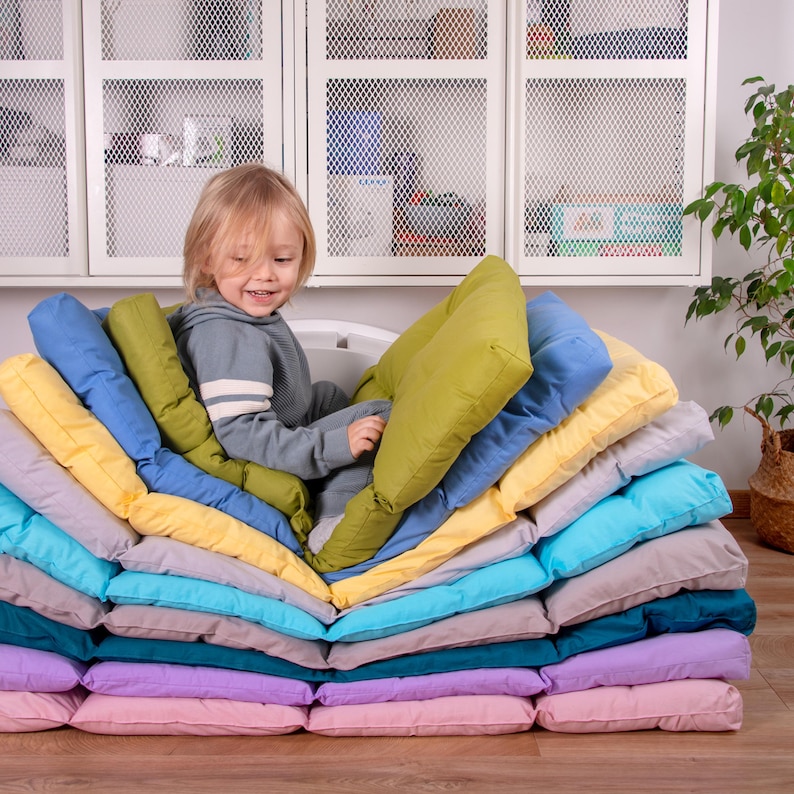 May include: A child sits on a stack of colorful cushions, arranged in a zig-zag pattern. The cushions are made of soft fabric and are perfect for creating a cozy and comfortable seating area.