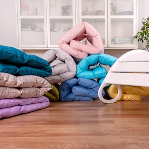 Può includere: Una varietà di cuscini colorati e piegati in diverse tonalità di blu, rosa, beige e giallo. I cuscini sono disposti a semicerchio su un pavimento di legno.