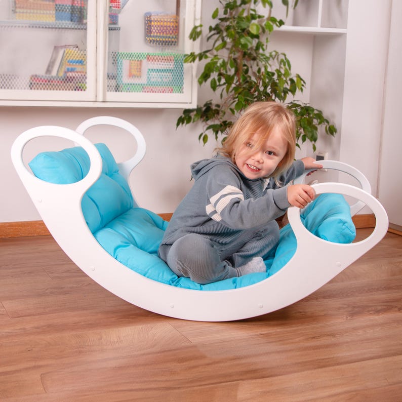 Op de afbeelding: Een witte houten schommelstoel met een blauw kussen. Een jong kind zit in de stoel en glimlacht.