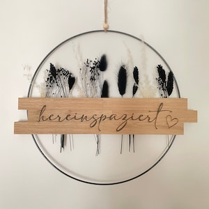Könnte beinhalten: Ein schwarzer Metallring mit getrockneten Blumen und Grün, der an einem Holzschild mit der Aufschrift "herein spaziert" mit einem kleinen Herzen hängt.