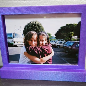 May include: A purple picture frame with a photo of two young girls hugging. The girls are smiling and the photo is set against a backdrop of a street scene with cars and trees.