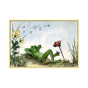 May include: A green frog with a pipe in its mouth is lying on its back in a field of grass. A brown hat with a pink flower is on a stick in the background. The text "Keepsake Fabrics" is visible in the lower left corner of the image.