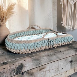 May include: An oval-shaped baby changing basket featuring a woven design in alternating bands of beige and sage green cord. The basket includes two integrated handles and a white mattress pad decorated with a pattern of small green eucalyptus leaves.