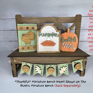 May include: A miniature wooden bench with fall-themed inserts. The inserts include a turkey, a pumpkin pie sign, and a pumpkin with the word "thankful." A banner with acorns and leaves is strung across the front. The bench is brown and the inserts have green, orange, and natural wood tones.