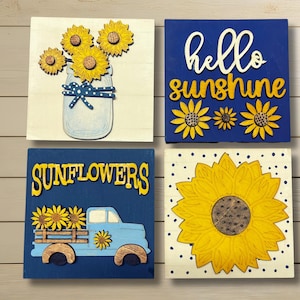 May include: Four square wooden signs with sunflower designs. One features sunflowers in a mason jar, another says "hello sunshine", a third shows a truck with sunflowers, and the last has a large sunflower.