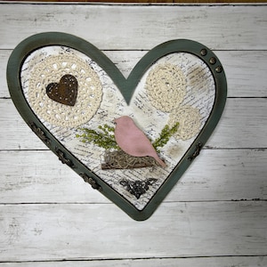 May include: A heart-shaped wall decor piece with a distressed green frame. Inside, a pink bird sits on a nest, surrounded by crochet doilies and a brown heart. The background features script writing.
