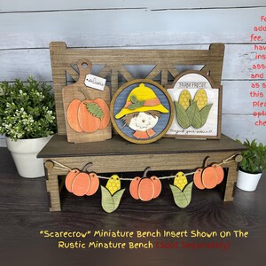 May include: A wooden miniature bench with fall-themed decorations. It features a pumpkin cutting board, a scarecrow, and a "Farm Fresh" sign with corn. A garland of pumpkins and corn adorns the front. The bench is brown, and the decorations are orange, yellow, and green.