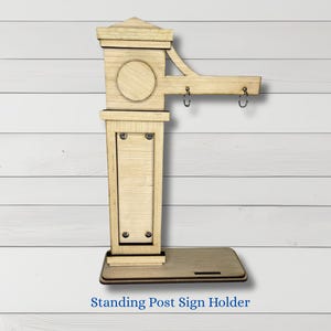 May include: A wooden standing post sign holder with a light brown finish. The sign holder has a rectangular base, a tall central post, and a horizontal arm with two hooks. The text "Standing Post Sign Holder" is at the bottom.