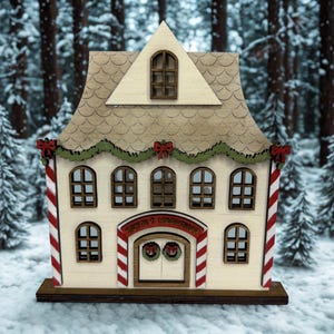 May include: A wooden Christmas house with a cream-colored facade, adorned with red and white candy cane stripes, green garland, and red bows. The house features multiple windows and a sign above the double doors. The background shows a snowy forest.