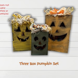 May include: A set of three wooden pumpkin-shaped boxes in varying sizes and colors. Each box features a carved jack-o'-lantern face and is adorned with fabric and straw accents. The text "Three Box Pumpkin Set" is displayed at the bottom.