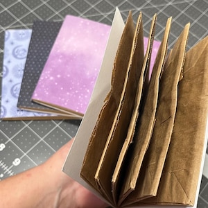 May include: Handmade journals with various patterned covers, including a light blue celestial design, a dark gray polka dot pattern, and a purple watercolor effect. The open journal reveals brown paper pages.