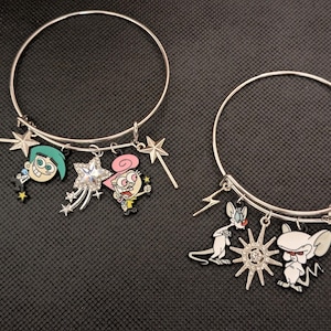 May include: Two silver-toned wire bangle bracelets featuring enamel character charms. The first bracelet includes Cosmo and Wanda figures with star wands and a crystal star. The second bracelet features Pinky and the Brain with a sunburst charm.