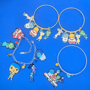 May include: Two gold-tone wire bangles and two silver-tone bracelets featuring colorful enamel charms of animated characters. The charms depict various figures in blue, yellow, red, and green, with some bracelets including small colored beads.