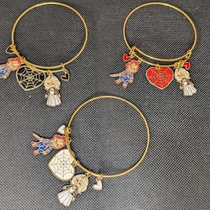 Op de afbeelding: Drie goudkleurige armbanden met bedels. Elke armband heeft een Chucky-poppenbedel, een hartvormige bedel met een spinnenwebontwerp en een poppenbedel. De bedels zijn rood, wit, zwart en blauw.