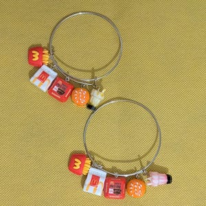 May include: Two silver bangle bracelets with food-themed charms. Charms include a hamburger, french fries, a milkshake, and McDonald's packaging. The charms are in red, white, orange, and yellow.