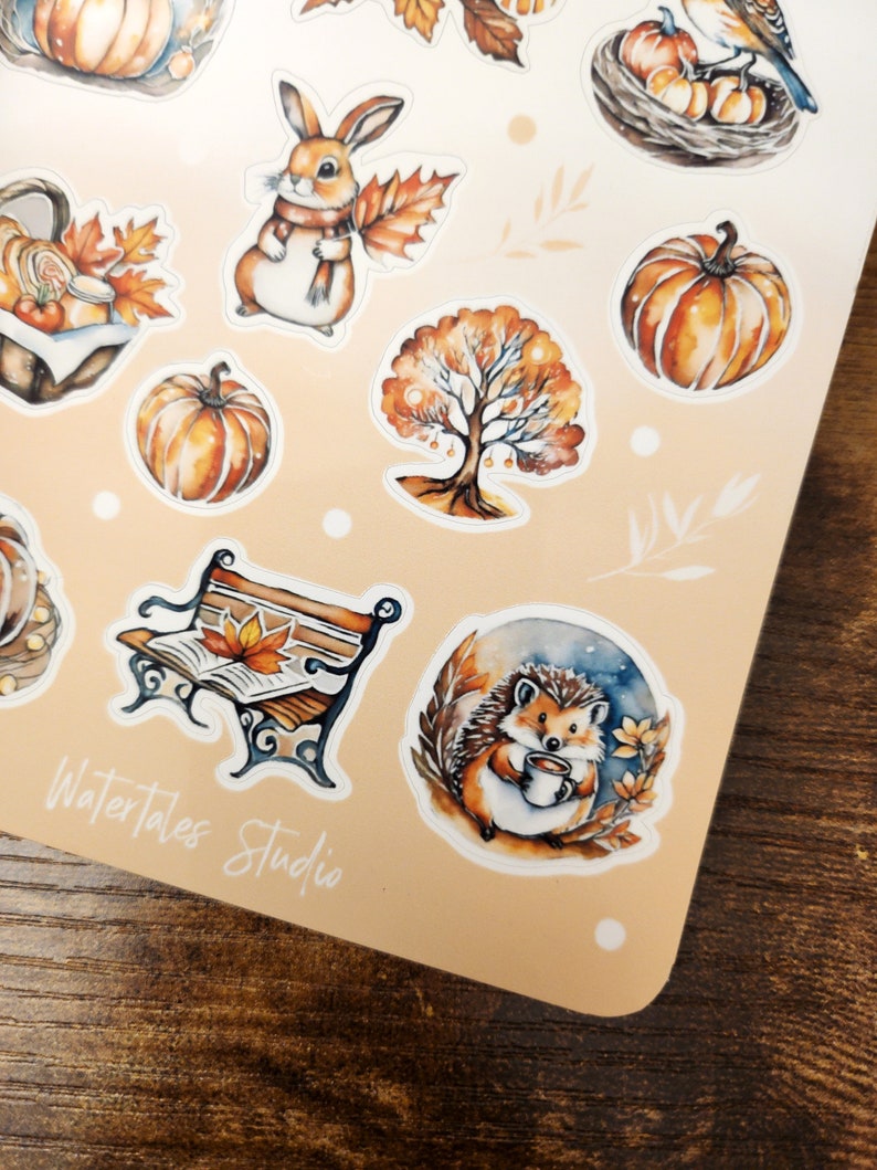 May include: A sheet of fall-themed stickers featuring watercolor illustrations. Designs include pumpkins, a rabbit with a scarf, a tree, a bench with a leaf, and a hedgehog holding a mug. The text "Watertales Studio" is visible.