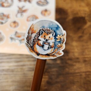 May include: A watercolor sticker of a hedgehog holding a mug of coffee, surrounded by autumn leaves and a blue and orange background. Additional stickers with fall themes are visible in the background.