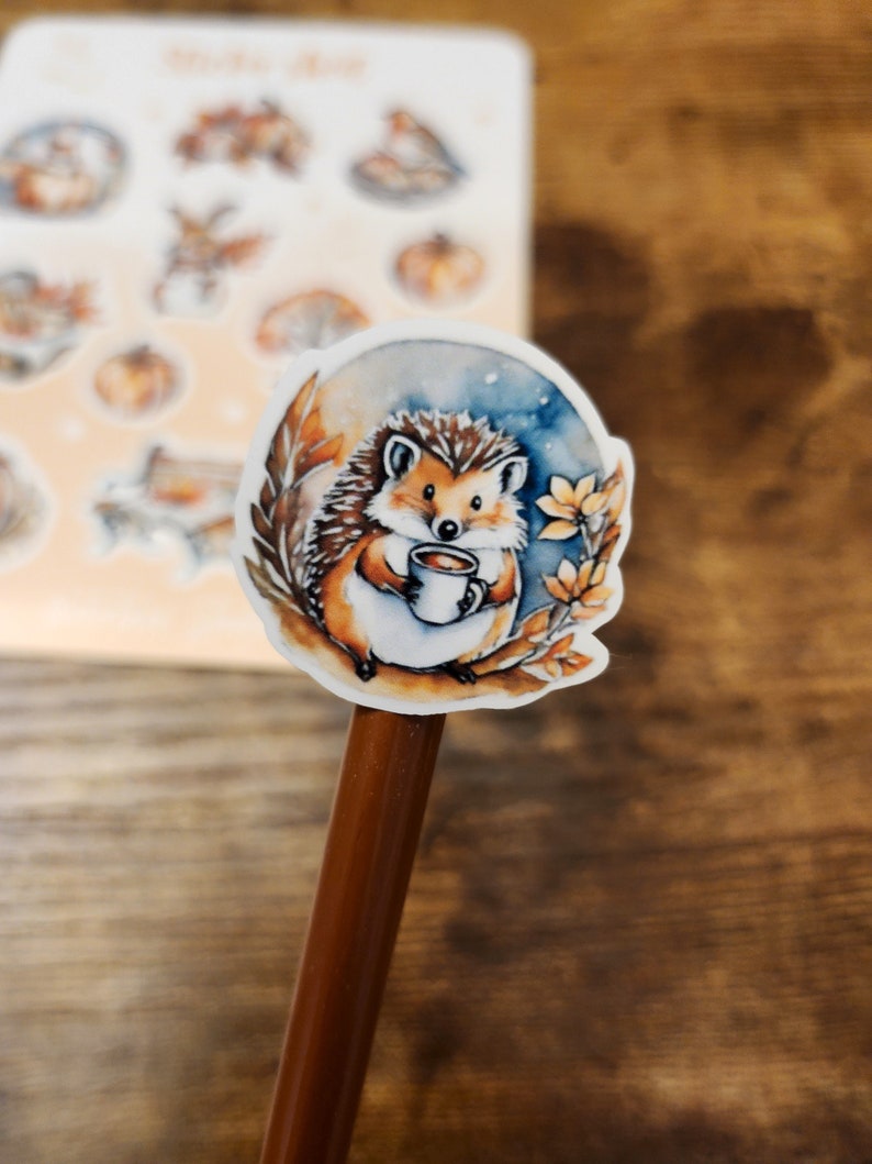 May include: A watercolor sticker of a hedgehog holding a mug of coffee, surrounded by autumn leaves and a blue and orange background. Additional stickers with fall themes are visible in the background.