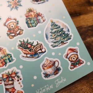 May include: A sheet of Christmas-themed stickers featuring watercolor illustrations. Designs include a decorated Christmas tree, teddy bears in Santa hats, wrapped gifts, a cake, a sleigh, a mug of hot chocolate, and a Christmas stocking. The text "Watertales Studio" is visible.