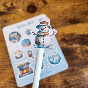 May include: A white pen topped with a snowman sticker, featuring a brown hat, scarf, and twig arms. A light blue sticker sheet with winter-themed illustrations is in the background. The text "Sticker sheet" is visible.