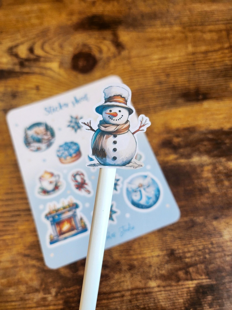 May include: A white pen topped with a snowman sticker, featuring a brown hat, scarf, and twig arms. A light blue sticker sheet with winter-themed illustrations is in the background. The text "Sticker sheet" is visible.
