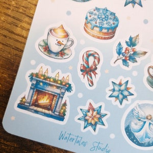 May include: A sheet of winter-themed stickers featuring illustrations of a snowman in a mug, a cake with snowflakes, a fireplace, and a candy cane. The stickers are in shades of blue, white, and gold. The text "Watertales Studio" is visible.