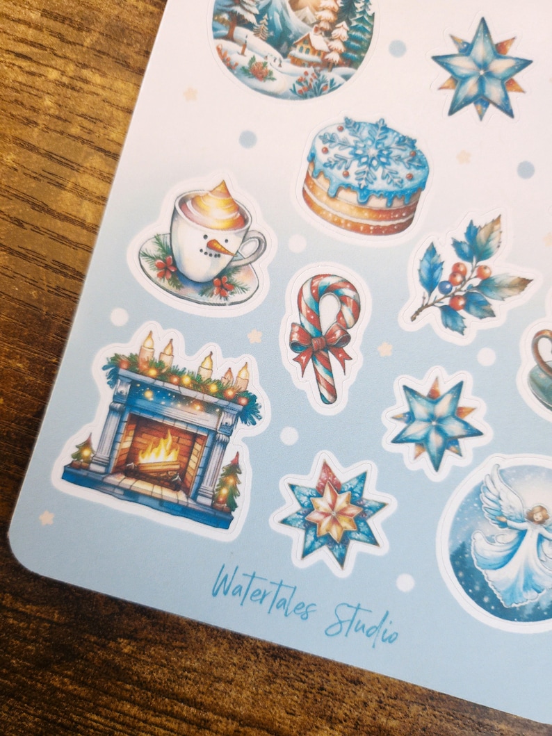 May include: A sheet of winter-themed stickers featuring illustrations of a snowman in a mug, a cake with snowflakes, a fireplace, and a candy cane. The stickers are in shades of blue, white, and gold. The text "Watertales Studio" is visible.