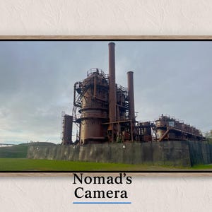 May include: Framed photograph of an industrial complex with tall smokestacks and a complex network of pipes and structures. The image is set against a cloudy sky and a green grassy area. The text "Nomad's Camera" is at the bottom.