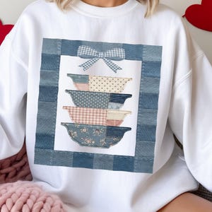 Peut inclure: Sweat-shirt blanc avec un motif patchwork de bols empilés dans divers motifs et couleurs, dont du denim bleu, du rose et des imprimés floraux. Un nœud vichy bleu et blanc est placé en haut du motif. Le motif est encadré d'une bordure en denim bleu.