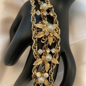 Puede incluir: Pulsera dorada con diseño de doble cadena. La pulsera está adornada con perlas de imitación, cristales transparentes y detalles florales. Una joya vintage.