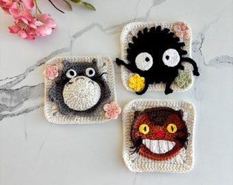 Pode incluir: Três bases de copos quadradas de crochê com personagens de um filme de animação popular. Uma base de copo retrata um Totoro cinza com flores rosa, outra um sprite de fuligem preto e a terceira um personagem felino marrom com um sorriso largo. As bases de copos estão em uma superfície de mármore branco.