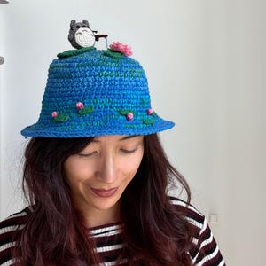 Peut inclure: Un chapeau bob en crochet bleu orné d'une petite figurine grise tenant une canne à pêche sur le dessus. Le chapeau est décoré de nénuphars verts et de petites fleurs roses dispersées sur la surface, créant une scène d'étang sur la calotte.
