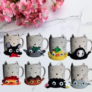 Può includere: Otto tazze in ceramica beige chiaro, ognuna con un design a forma di muso di gatto con orecchie, baffi e naso. Ogni tazza è decorata con una copertura all'uncinetto colorata e unica attorno alla base, raffigurante vari personaggi.