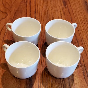 May include: Four white ceramic cups with handles are arranged on a wooden surface. The cups have a simple, rounded design and a smooth, glossy finish. The cups are empty and ready to be filled.