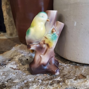 May include: A ceramic figurine of a small bird with yellow and green plumage perched on a brown tree stump. The bird has a light-colored beak and is positioned on the left side of the textured, branch-like structure, which features a hollow opening at the top.