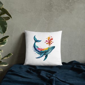 May include: White square pillow featuring a watercolor illustration of a whale. The whale is depicted in shades of blue, teal, yellow, and red, with a colorful spray emanating from its blowhole. The pillow is placed on a dark blue surface.