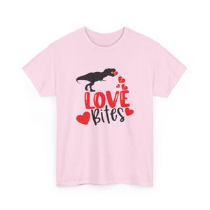 May include: A light pink t-shirt featuring a black Tyrannosaurus Rex silhouette holding hearts. The words "LOVE Bites" are in red and black, with red heart accents. A fun Valentine's Day design.