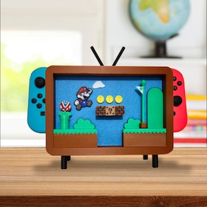 May include: A brown framed shadow box depicting a Super Mario Bros. scene on a blue backdrop. The scene includes Mario, coins, a question mark block, and a green pipe. Two game controllers are attached to the sides of the frame.