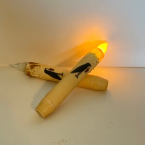 May include: Two battery-operated candles with a black and red design of a bird and a star. The candles are lit and glowing orange.