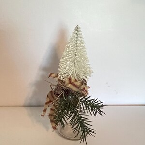 May include: A small, clear glass jar decorated with a miniature white Christmas tree, sprigs of evergreen, and a red and cream striped ribbon. The jar is sitting on a white surface.