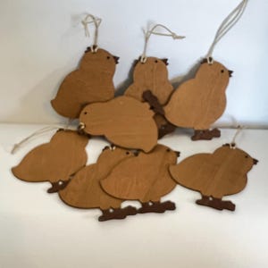 May include: A collection of wooden bird ornaments. Each bird is cut from light brown wood, featuring a simple bird shape with small feet and a string for hanging. The birds are clustered together, suggesting a decorative or craft theme.
