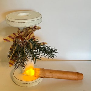 May include: White wooden candle holder with a rustic, distressed finish. The holder features a decorative burlap and ribbon bow with a pine sprig. A battery-operated candle is placed in the holder.