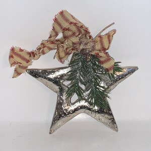 May include: A silver star-shaped ornament with a rustic, textured finish. It is decorated with a sprig of green pine and a red and white striped ribbon.