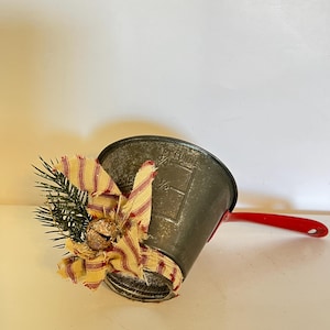 May include: A small, vintage metal bucket with a red handle, decorated with a bow of red and cream striped fabric, a small bell, and sprigs of evergreen. The bucket has a distressed finish.