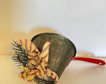 May include: A small, vintage metal bucket with a red handle, decorated with a bow of red and cream striped fabric, a small bell, and sprigs of evergreen. The bucket has a distressed finish.