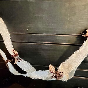 May include: A decorative garland featuring a long, off-white frayed fabric ribbon draped in a loop. The ribbon is accented with small, dark brown and tan patterned fabric bows tied at intervals along the length of the textured, fringed material.