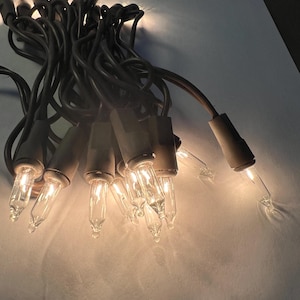 May include: A string of clear incandescent mini lights with dark brown wire and sockets. The lights are bundled together in a cluster, with several individual bulbs illuminated, casting a warm glow from the clear glass filaments.