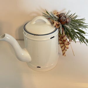 May include: White ceramic teapot with a distressed finish, decorated with a sprig of faux pine, red and white plaid ribbon, and brown pine cones.
